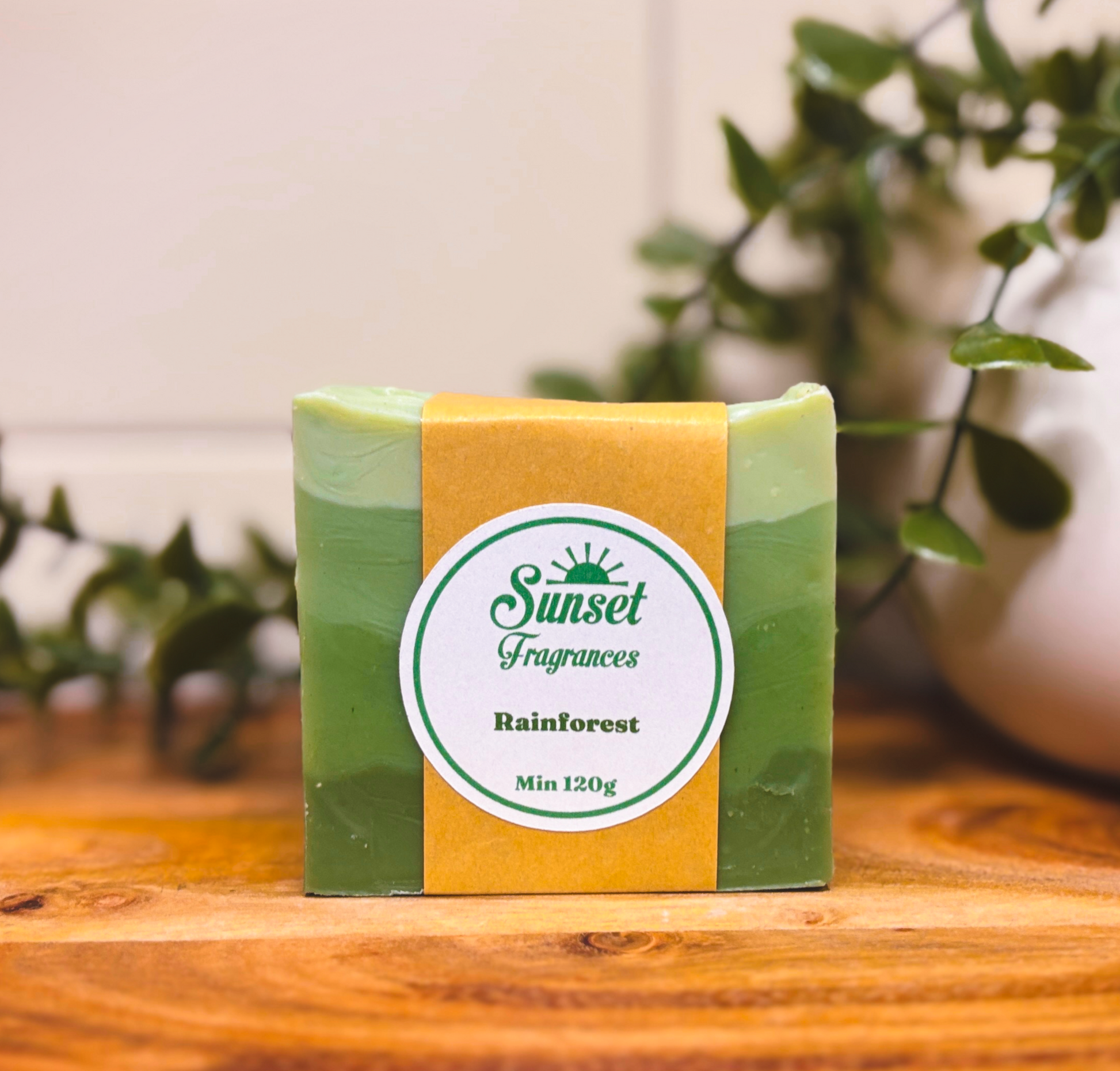 Bar of soap with a 'Sunset Fragrances' label on a wooden surface with plants in the background