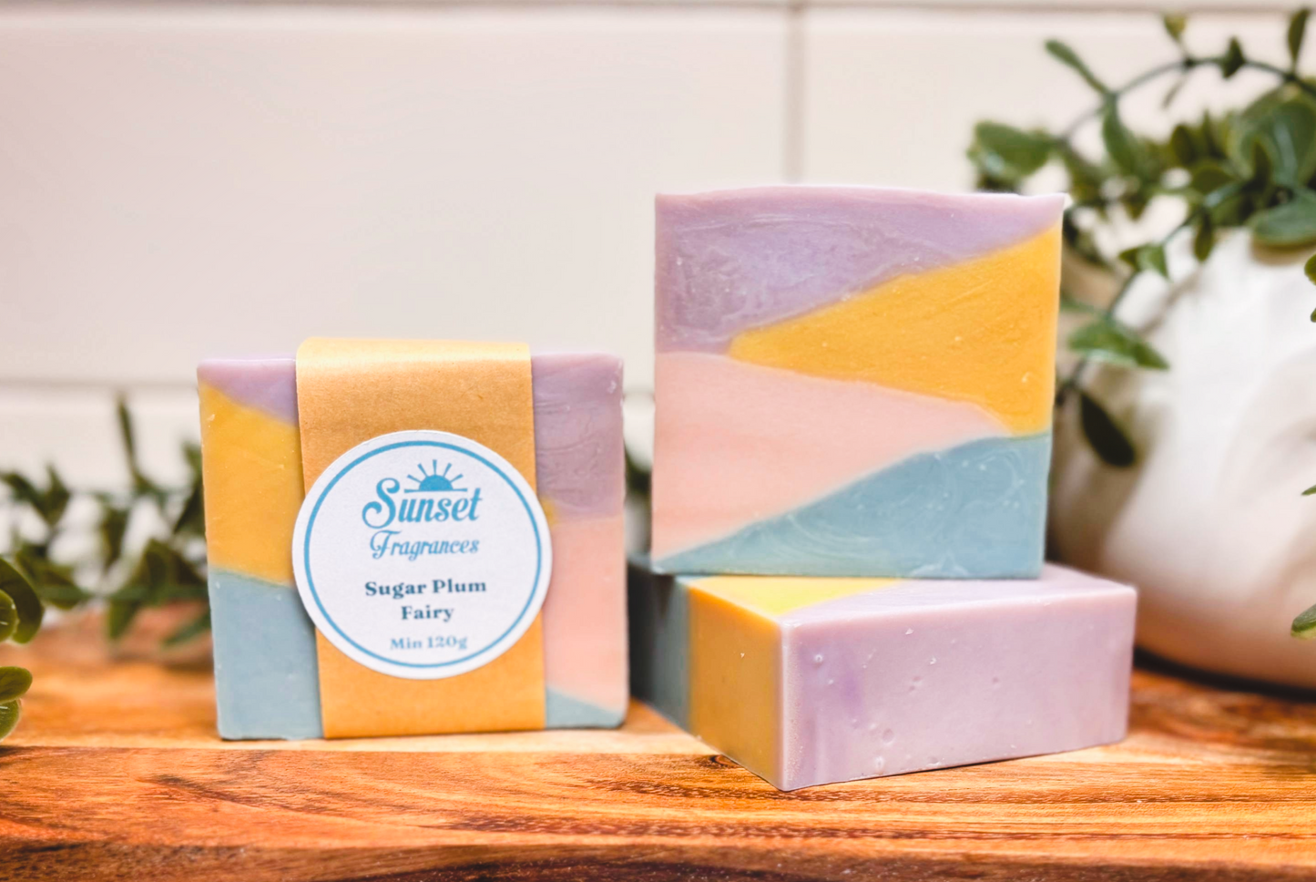 Three colorful soap bars on a wooden surface with a plant in the background